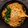 Egg noodles getting tossed in peanut sauce with chopped up peanuts, cilantro and onions getting mixed in a cast iron pan. 