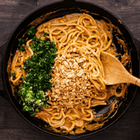 Egg noodles getting tossed in peanut sauce with chopped up peanuts, cilantro and onions getting mixed in a cast iron pan. 
