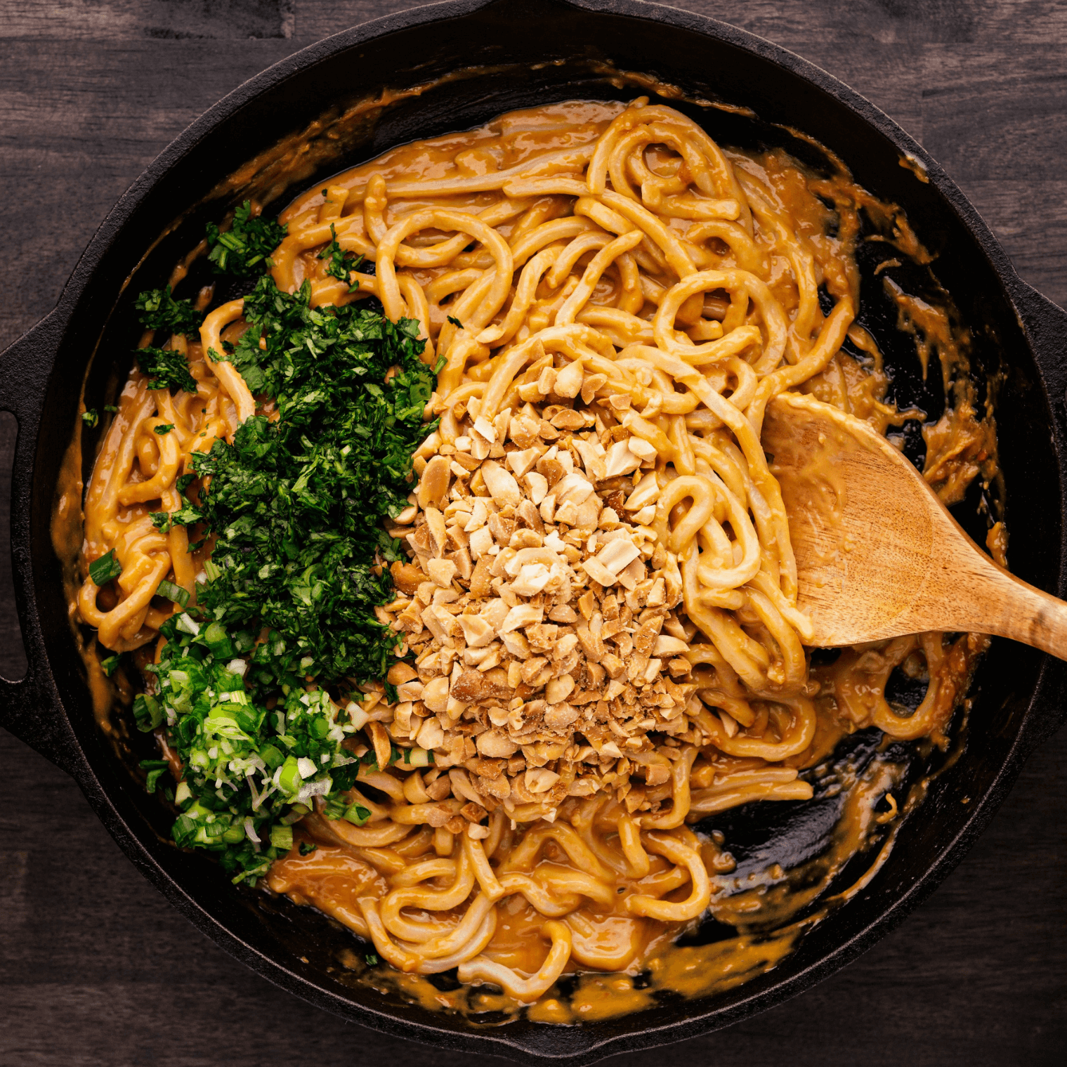 Egg noodles getting tossed in peanut sauce with chopped up peanuts, cilantro and onions getting mixed in a cast iron pan. 