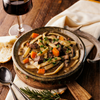 Wide egg noodles in broth of stew plated on rustic table with wine in background and bread. 